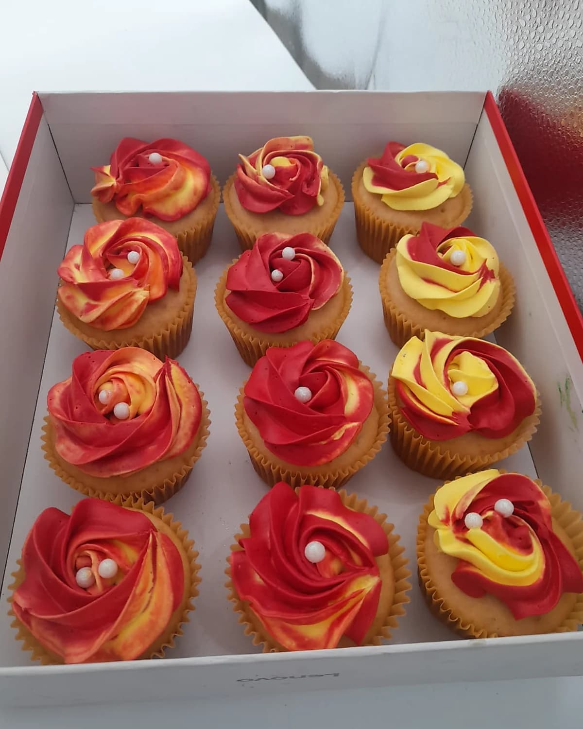 Caja de doce cupcakes de vainilla con rosetas de buttercream en rojo y amarillo, decorados con perlas blancas